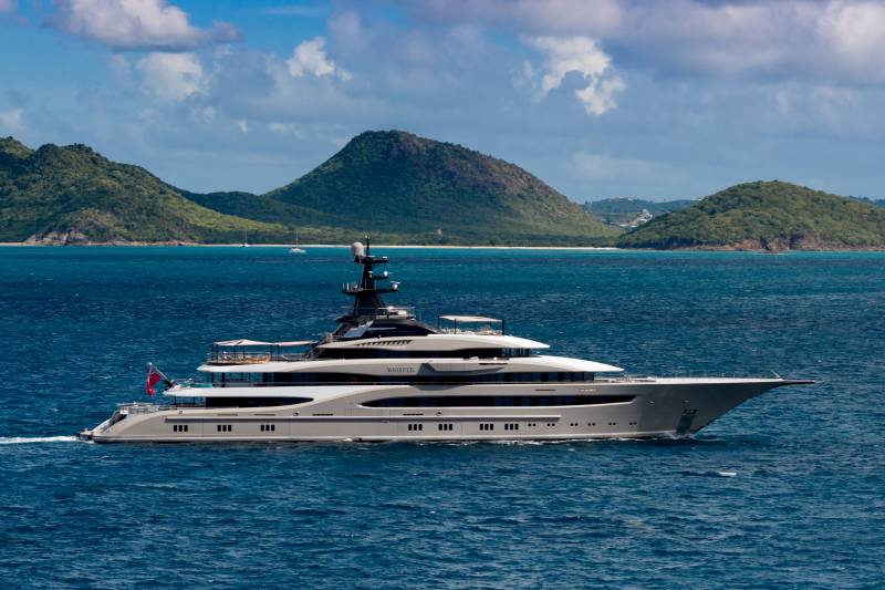 A luxurious superyacht glides through the turquoise waters of a tropical bay, with lush green hills in the background.
