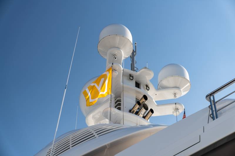 A bright yellow flag with white geometric symbols flutters against a clear blue sky atop a sleek white yacht.
