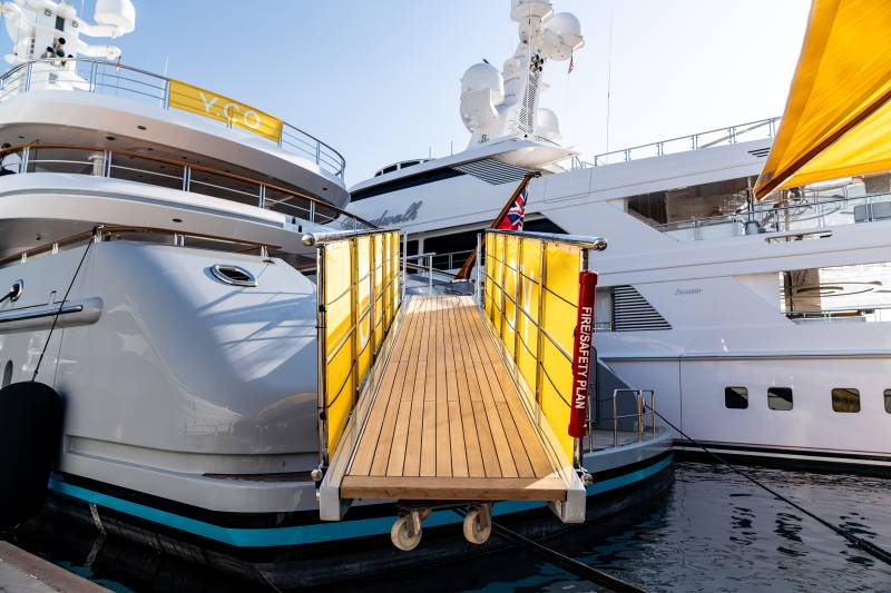 A wooden gangway with yellow safety banners leads onto a luxurious yacht docked in a harbor.