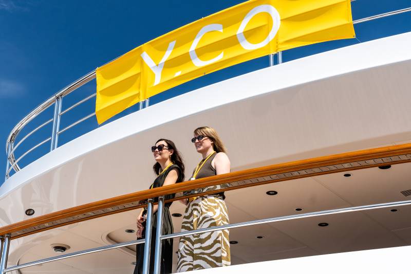 Two women in sunglasses stand on a yacht deck under a yellow Y.CO banner, possibly at the Palm Beach International Boat Show.
