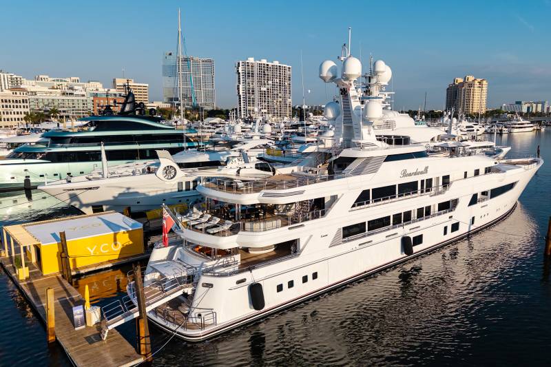 Luxurious yachts, including the "Boardwalk," are docked in a sunny harbor with modern buildings in the background.