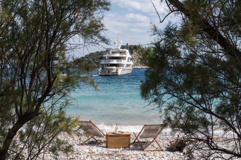 Relax on a pebble beach with two lounge chairs and a bottle of wine, overlooking a luxurious yacht anchored in the turquoise bay.