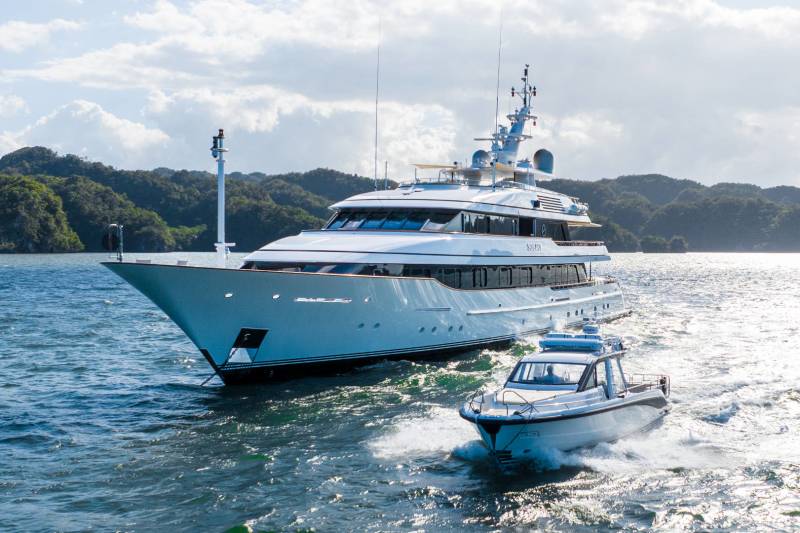 A sleek superyacht cruises alongside a smaller tender boat in a tranquil bay surrounded by lush green hills.