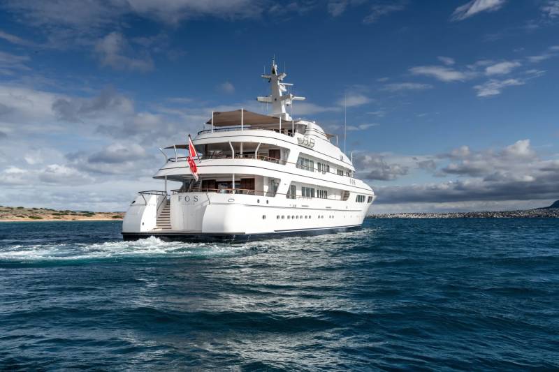 A large, luxurious white yacht is anchored in the calm, turquoise waters of a scenic coastal area, with a clear blue sky and fluffy white clouds in the background.