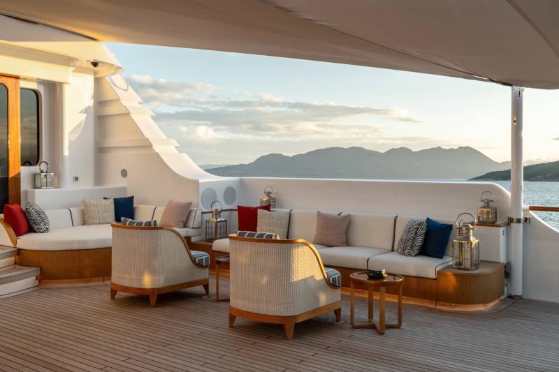 A cozy outdoor lounge area on a yacht or cruise ship, with comfortable seating and a stunning view of mountainous islands in the background.