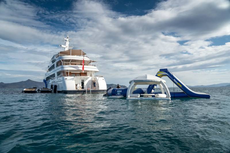 A large luxury yacht is anchored in the ocean, with inflatable water slides and other recreational equipment visible in the foreground against a backdrop of cloudy blue skies.