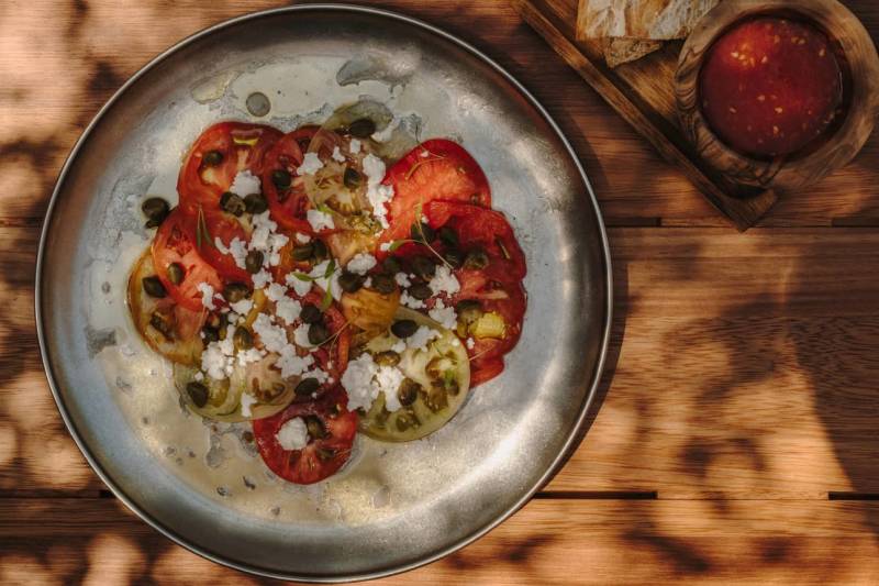 A gray ceramic dish filled with roasted red bell peppers, crumbled feta cheese, and herbs sits on a wooden surface, with a whole red bell pepper visible in the background.