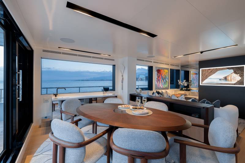 A modern and stylish dining area with a large oval wooden table surrounded by comfortable chairs, overlooking a scenic body of water through the large windows in the background.