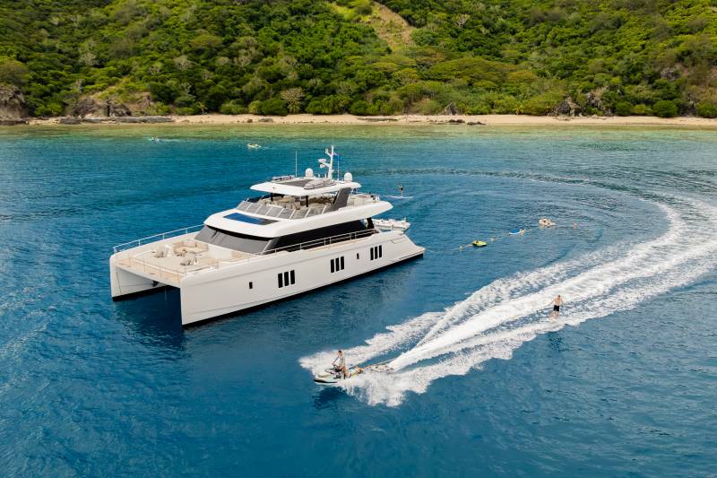 A sleek catamaran yacht floats on calm blue water in the foreground, while a person on a jet ski creates a white wake, and a lush green, tree-covered island rises in the background.