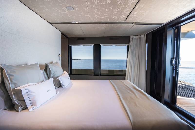 A neatly made bed with plush pillows occupies the foreground, while large windows in the background reveal a serene ocean view.