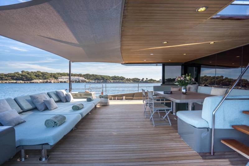 A spacious yacht deck with lounge chairs and a dining table in the foreground overlooks a calm sea and a tree-lined coast in the background.