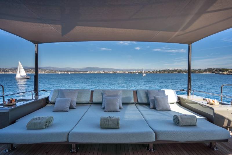 A spacious sun deck with plush cushions and rolled towels faces a tranquil blue sea dotted with sailboats and a distant, sun-drenched coastline.