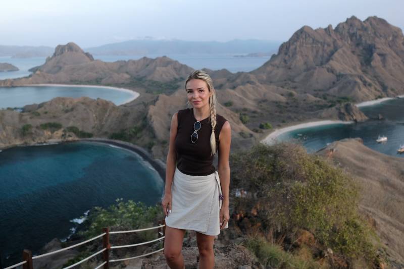 A blonde woman stands on a grassy hilltop in the foreground, with a stunning vista of rugged islands and turquoise bays in the background.