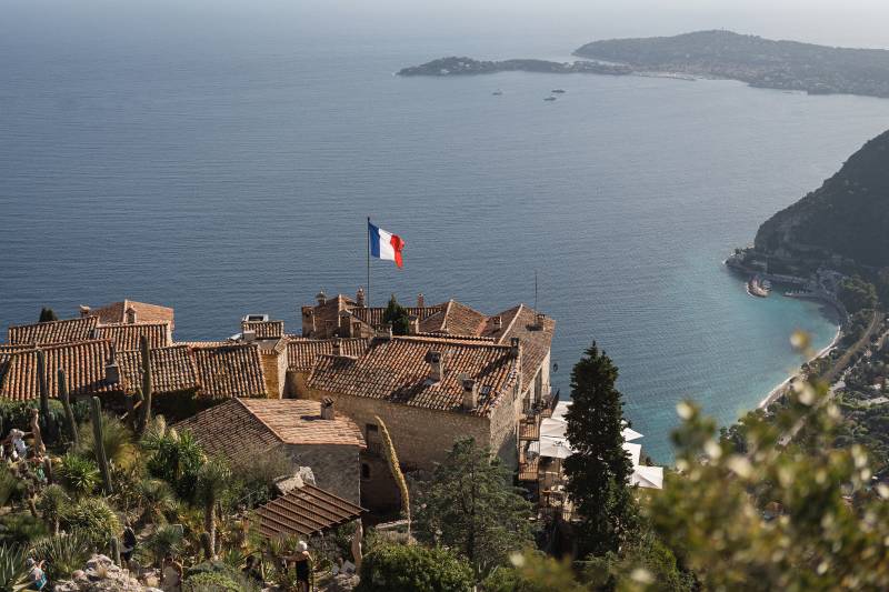 A French flag flies over a village with terracotta roofs in the foreground, overlooking a sparkling blue bay and distant coastline in the background.