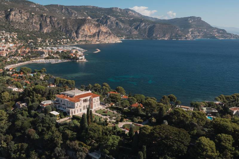 A grand villa with manicured gardens sits nestled amongst lush greenery in the foreground, overlooking a sparkling blue bay dotted with sailboats and a coastal town nestled against dramatic cliffs in the background.