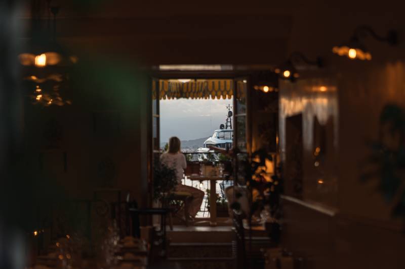 A woman sits at a table in a dimly lit room, looking out at a large white yacht docked in a harbor.