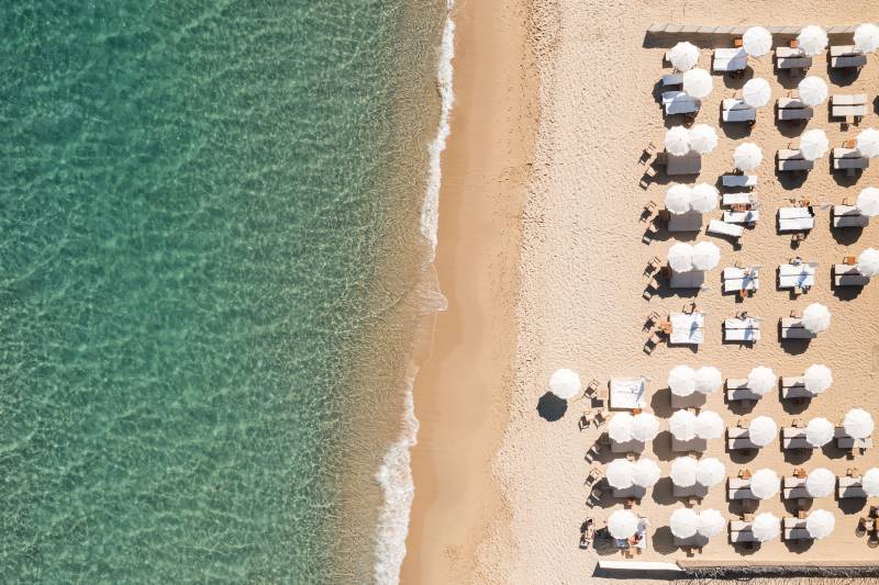 Crystal clear turquoise water laps onto a sandy beach lined with rows of white umbrellas and lounge chairs.