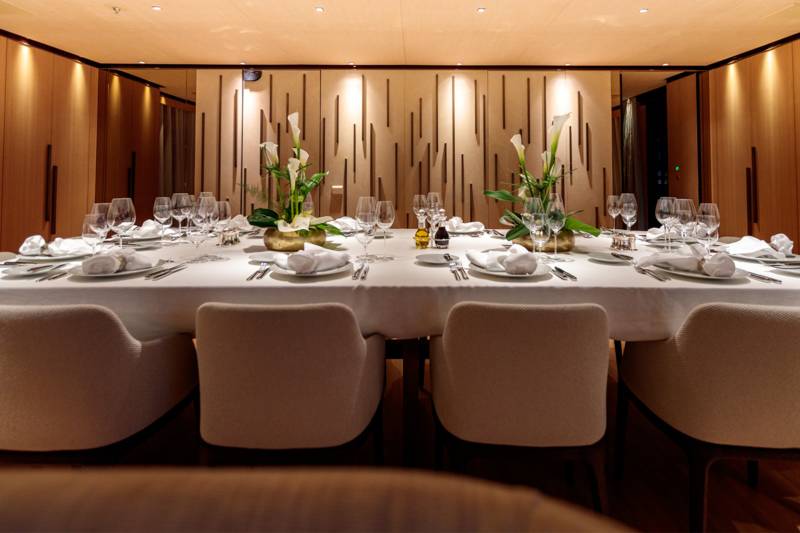 A long, elegantly set dining table with white chairs in the foreground is surrounded by warm, wood-paneled walls with decorative lighting in the background.