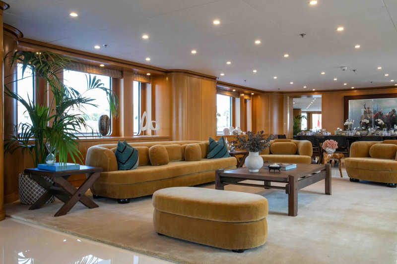 A plush mustard-colored sofa and ottoman dominate the foreground of a luxurious yacht interior, while the background reveals a dining area with a large painting.