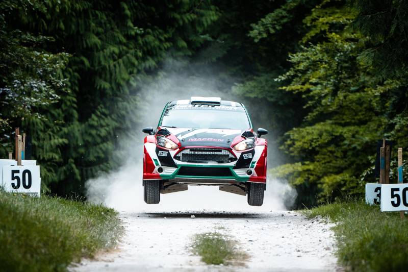 A rally car soars through the air, kicking up dust on a dirt track lined with trees and marked with "50" signs.