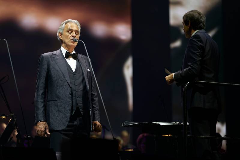 A man in a tuxedo sings into a microphone in the foreground, while a conductor stands behind him in the background.