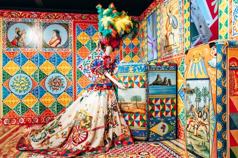 A mannequin in a voluminous, patterned gown stands amidst a vibrant, tiled room adorned with colorful artwork and decorative panels.