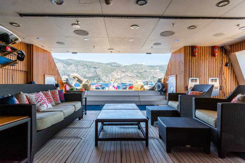 A luxurious outdoor lounge area with wicker furniture and colorful cushions is set against a backdrop of a scenic coastal town and mountains.