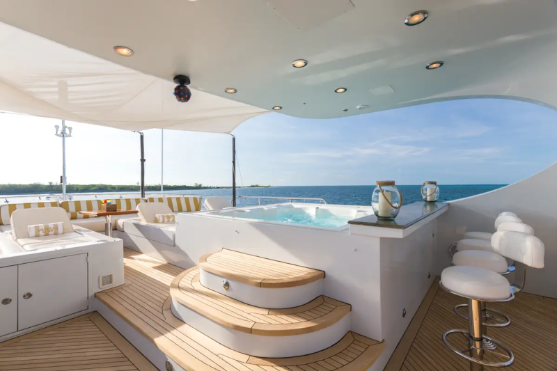 A luxurious yacht's deck features a bubbling hot tub and bar stools in the foreground, with a calm blue ocean and clear sky in the background.
