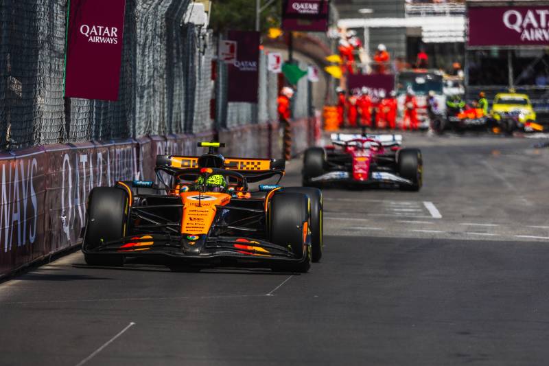 An orange and black Formula 1 car races down a track in the foreground, with a red Formula 1 car and pit crew visible in the blurred background.