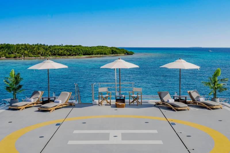 Lounge chairs and umbrellas are set up on a helipad overlooking a tropical ocean with a lush green island in the distance.