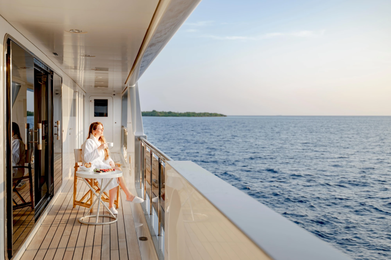 A woman in a white robe enjoys a drink on a yacht's deck, with the calm ocean and a distant island filling the background.