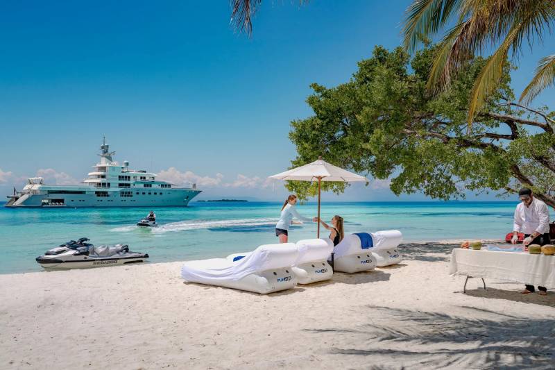 In the foreground, inflatable loungers and a table with refreshments sit on a sandy beach, while a large yacht cruises in the turquoise ocean under a clear blue sky in the background.