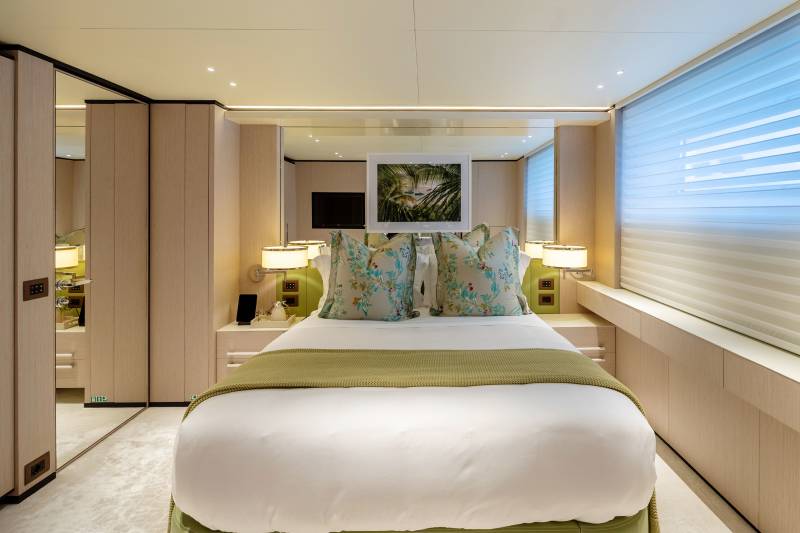 A neatly made bed with floral pillows is in the foreground, while mirrored closets and a large window with blinds fill the background of this luxurious cabin.