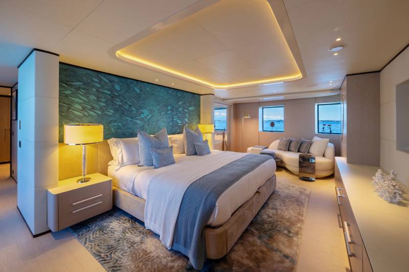 A luxurious yacht bedroom with a large bed in the foreground and a seating area with windows overlooking the water in the background.