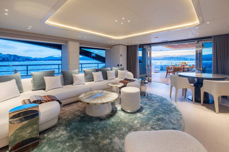 A luxurious yacht lounge with a large white sectional sofa and modern coffee tables in the foreground, opens to a deck with dining furniture and a scenic ocean view in the background.