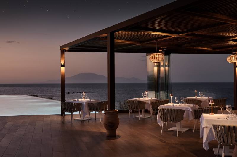 A romantic outdoor restaurant with tables set for dining is situated on a wooden deck overlooking the ocean and distant mountains under a starry night sky.
