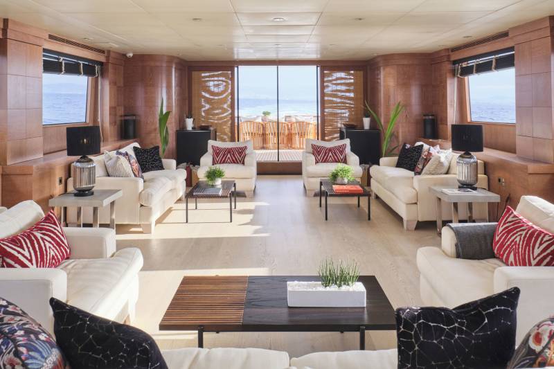 Plush white sofas with decorative pillows fill the foreground of a luxurious yacht lounge, while a bright beach scene is visible through large windows in the background.