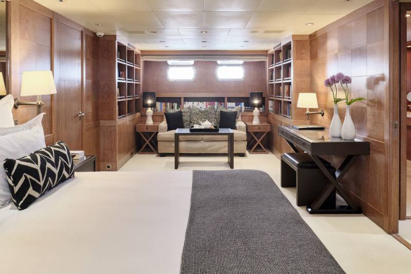 A luxurious bedroom with a large bed in the foreground and a desk and seating area in the background, all surrounded by wooden paneling and bookshelves.