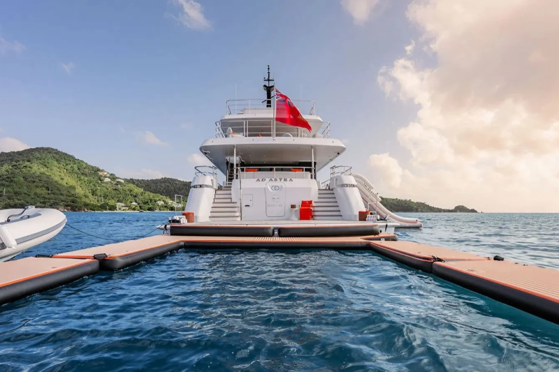 A large white yacht with a red flag is docked at a floating pier, with lush green islands and a cloudy sky in the background.