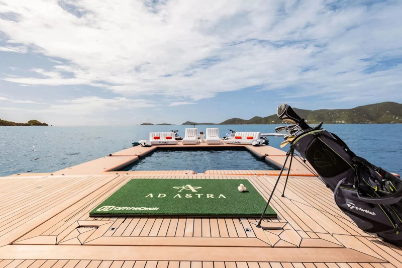 A golf bag and mat sit on a wooden deck in the foreground, with a pool and lounge chairs on a floating platform in the background, all set against a backdrop of the ocean and distant islands under a cloudy sky.
