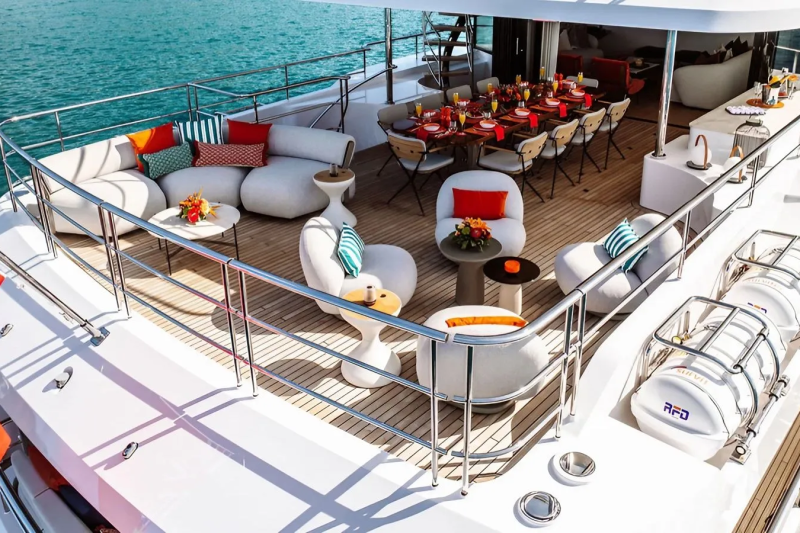 A luxurious yacht deck with comfortable seating and a dining table in the foreground, set against a backdrop of calm blue water.
