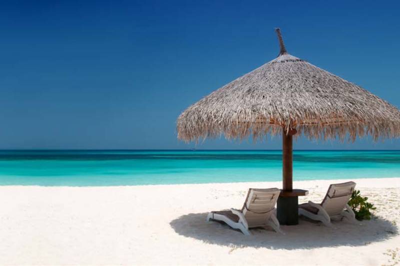 Two lounge chairs sit under a thatched umbrella on a white sand beach, with the turquoise ocean stretching to the horizon under a clear blue sky.