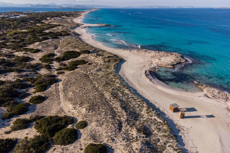 Sandy dunes dotted with scrub brush lead to a pristine white beach and turquoise waters, with a distant coastline under a clear blue sky.
