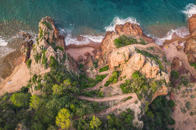 A winding path leads through lush green vegetation and rocky outcrops to a sandy beach, with the turquoise ocean waves crashing against the shore in the background.