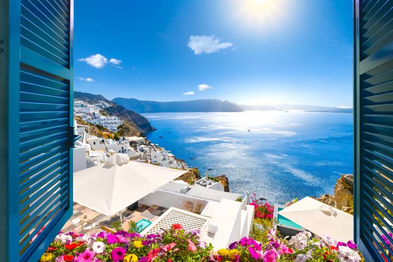 Brightly colored flowers in the foreground frame a sun-drenched view of white buildings cascading down a cliffside to a sparkling blue sea under a clear sky.
