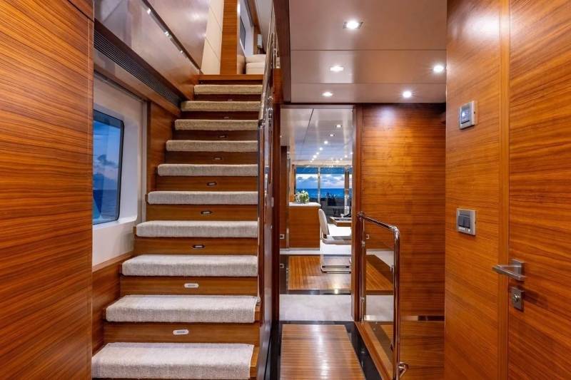 A luxurious yacht interior features a carpeted staircase in the foreground and a glimpse of a sunlit deck with ocean views in the background.