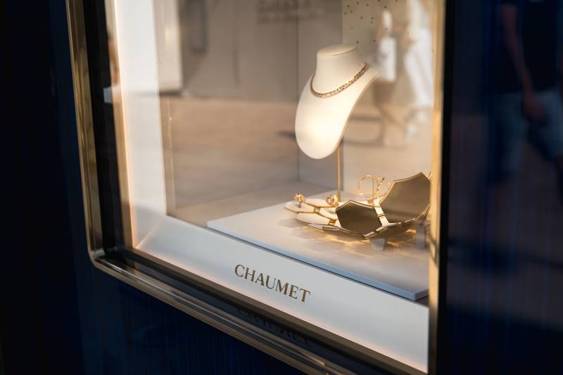 A diamond necklace is displayed on a white bust in a jewelry store window, with a blurred background of people walking by.