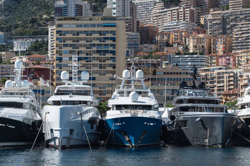 Several large yachts are docked in a harbor with a densely populated city built on a hillside in the background.