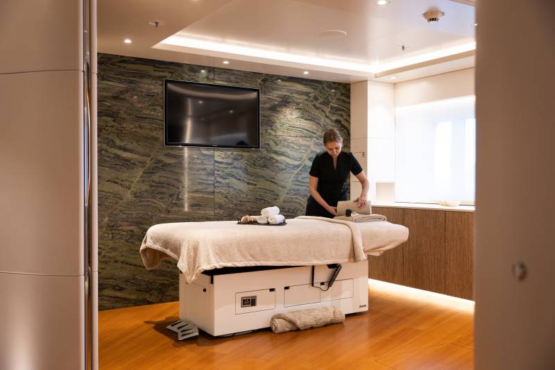 A massage therapist prepares a massage table in a spa room with a marble accent wall and a television in the background.