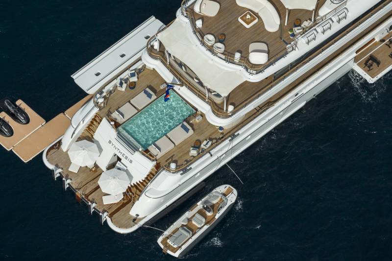 A luxurious yacht with a pool and lounge area is anchored in the deep blue sea, with a smaller tender boat nearby.